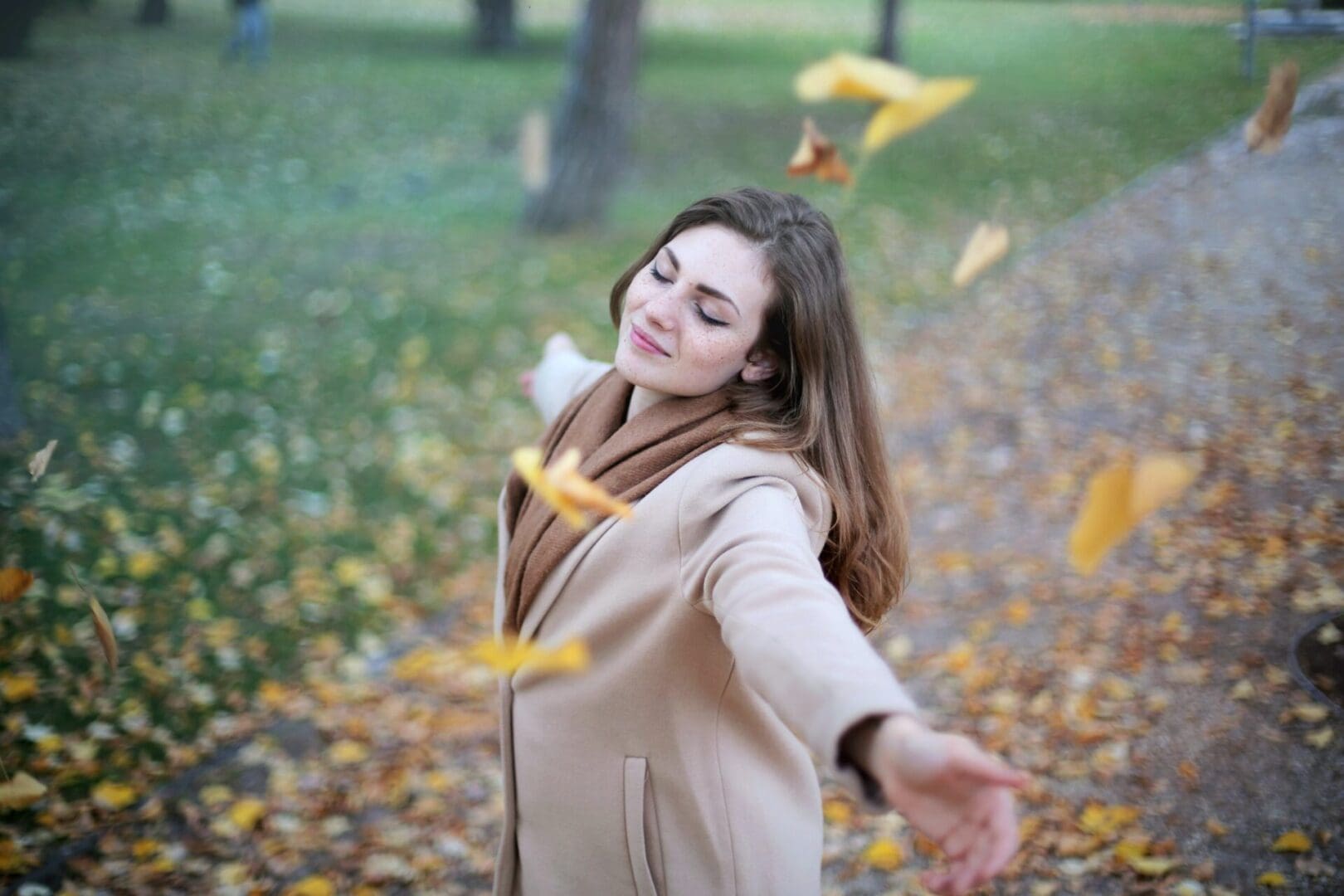 A woman in the park throwing leaves into the air.